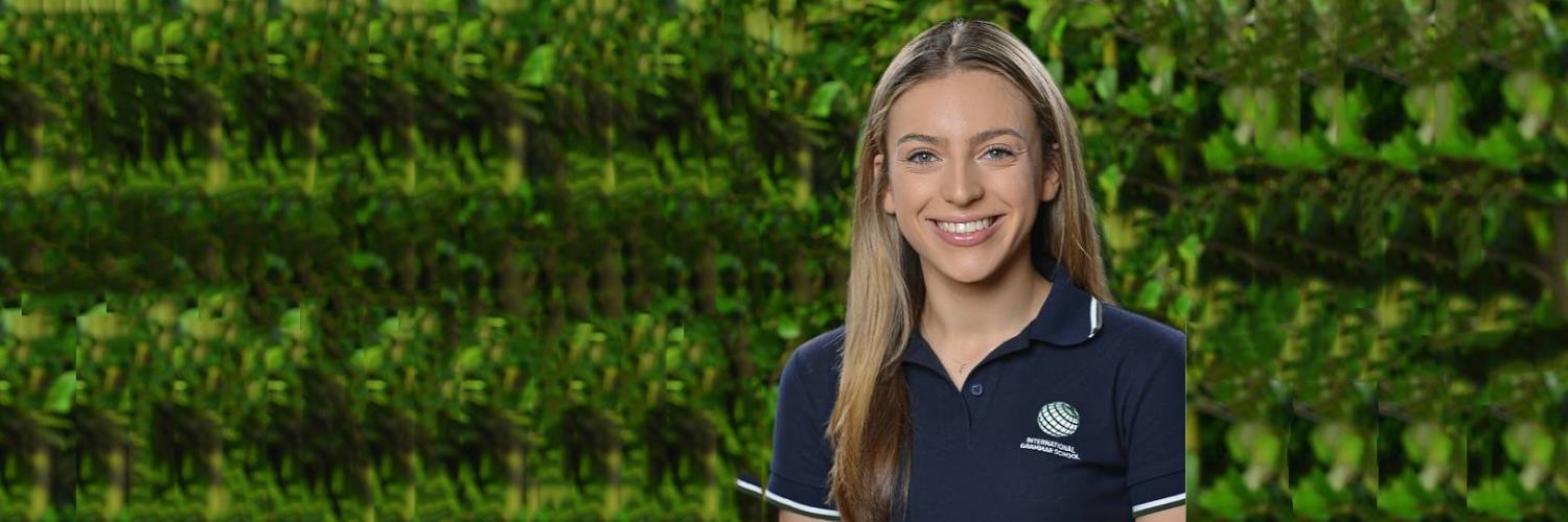 Catherine, a young woman with long blonde hair, wearing a navy blue collared shirt, smiles in front of a lush green leafy background that reflects the beauty of the State.