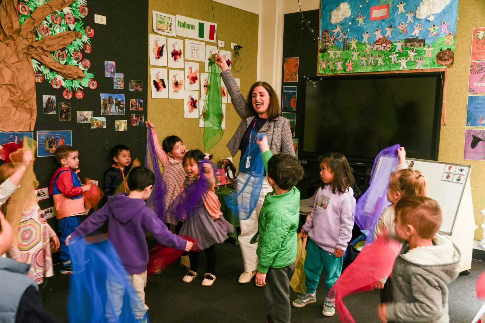 A teacher enthusiastically leads a group of young children in a classroom activity; the kids wave colourful scarves whilst smiling and moving to music. The classroom walls, filled with art and educational posters, highlight the power of music in early learning.