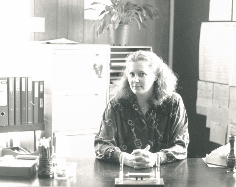 A woman with light-coloured hair sits at a desk in an office, hands clasped. Behind her are a filing cabinet and shelves with files, evoking old memories. A potted plant and papers pinned to the wall complete this black and white scene.