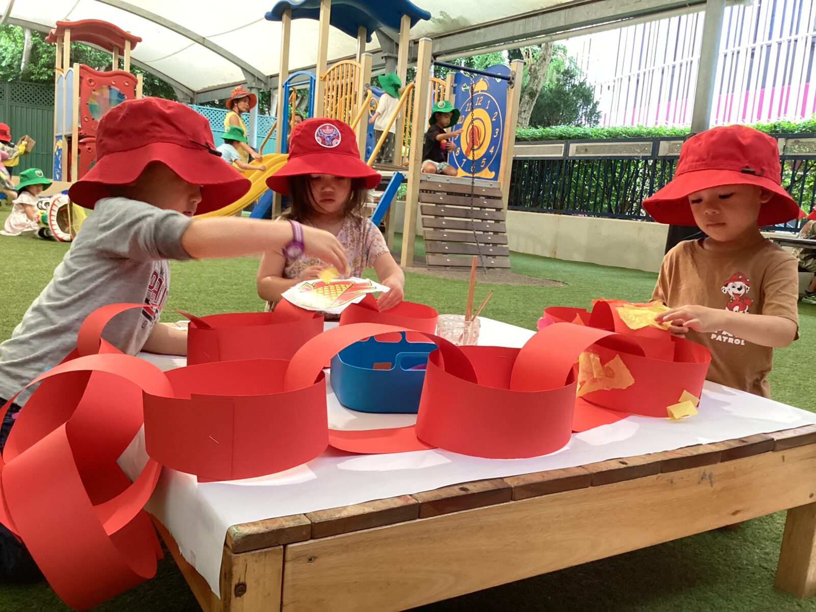Three young children wearing red hats sit at a table outdoors, making a long red paper chain for Chinese New Year. Play structures and more children are visible in the background, with greenery and an ELC play area surrounding them.