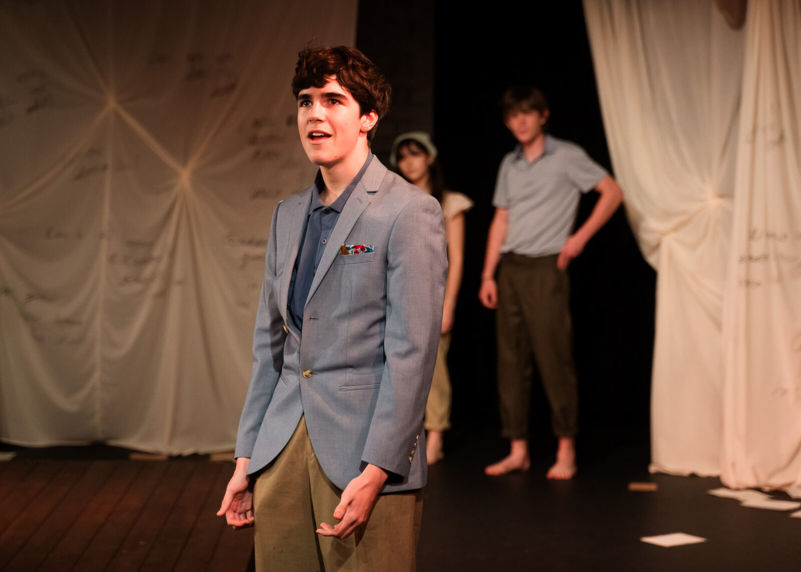A young person in a light blue blazer stands under stage lights, speaking with emotion in a Shakespeare play. Two other barefoot performers in casual clothes stand in the background near draped, script-covered fabric.