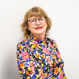 Shirley Alexander, a woman with short light brown hair and glasses, smiles at the camera. She is wearing a colourful floral blouse and stands against a plain white background.