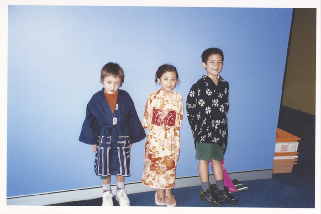 Three children stand in front of a blue wall, smiling at the camera. Two wear traditional Japanese-style clothing, whilst one wears a dark patterned outfit with shorts and trainers, reflecting harmony and diversity in their styles.
