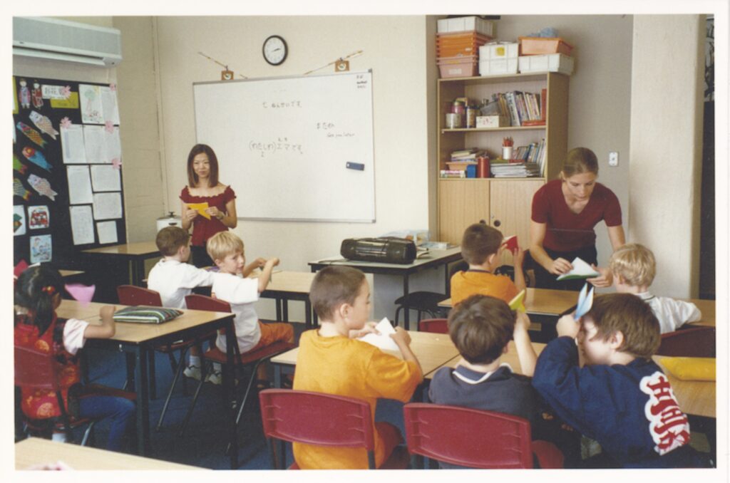 A classroom filled with young children of diverse backgrounds sits at desks, folding paper aeroplanes, while two teachers supervise. One stands by the whiteboard; the other leans over to help, creating an atmosphere of harmony among drawings and books on shelves and walls.