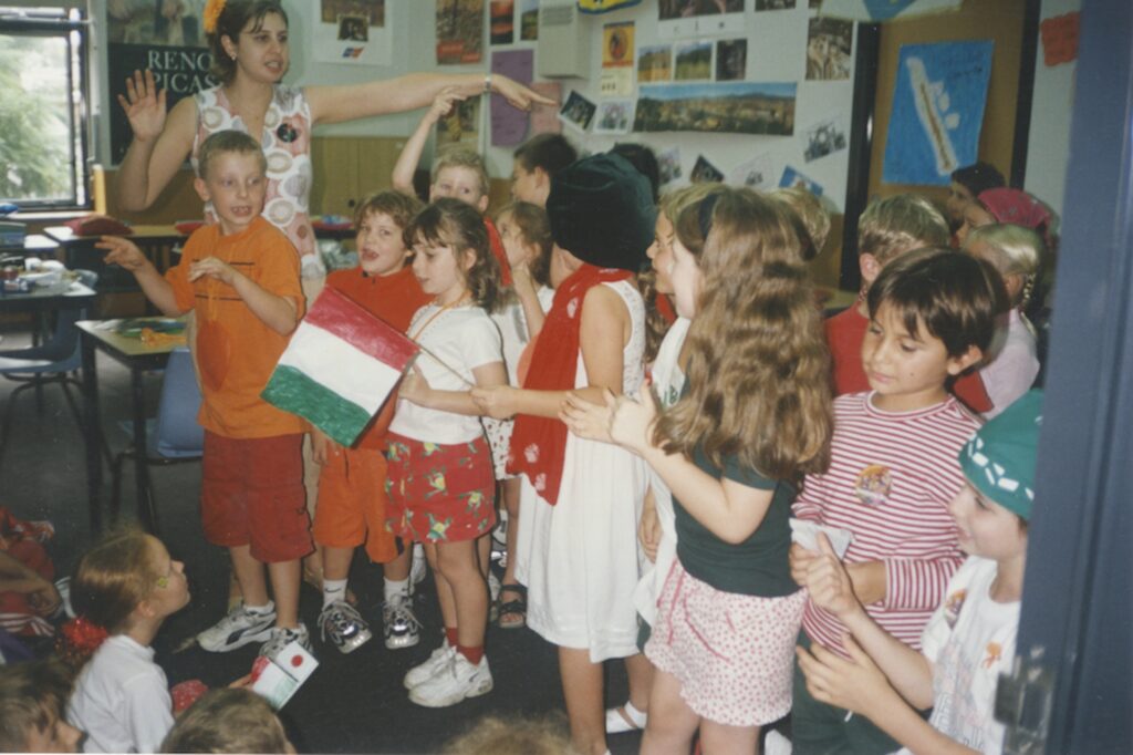 A group of young children in colourful clothes stand in a lively classroom, celebrating diversity as they hold or wear items with red, white, and green stripes. The walls are adorned with posters and photos whilst one adult gestures animatedly.