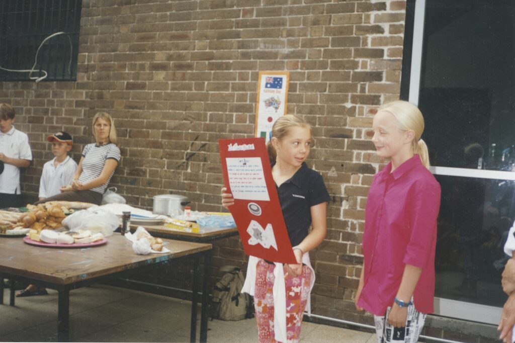 Two girls stand in front of a brick wall; one holds a red poster and looks at the other. A table with food and drinks is in the background, as people gather. Both girls are smiling, radiating harmony and celebrating diversity together.