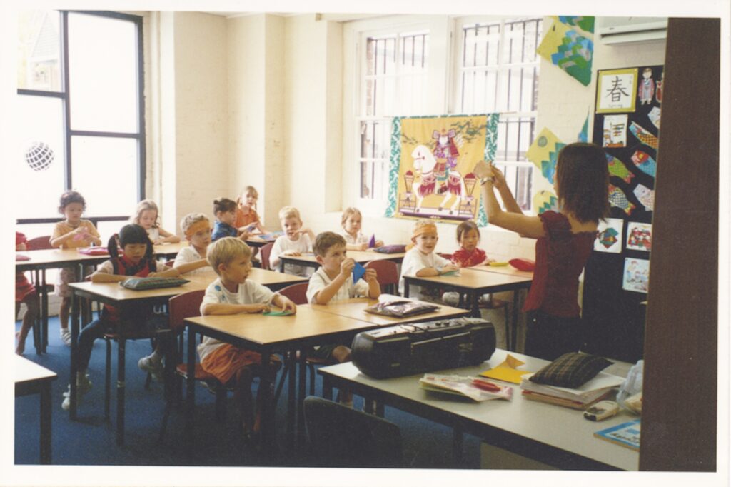 A teacher stands at the front of a brightly lit classroom, holding up an object for a diverse group of young children sitting at desks. Colourful artwork and posters decorate the walls, reflecting harmony as the children appear attentive and engaged.