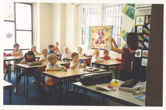A teacher stands at the front of a brightly lit classroom, holding up an object for a diverse group of young children sitting at desks. Colourful artwork and posters decorate the walls, reflecting harmony as the children appear attentive and engaged.