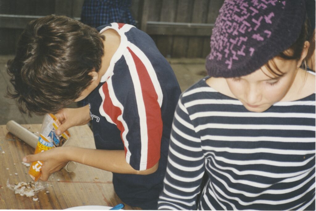 Two children stand at a wooden table preparing food in harmony. One, in a striped shirt and hat, looks down, whilst the other, in a sporty shirt, squeezes cheese from a packet. A rolling pin and chopped ingredients add to the scene’s diversity.