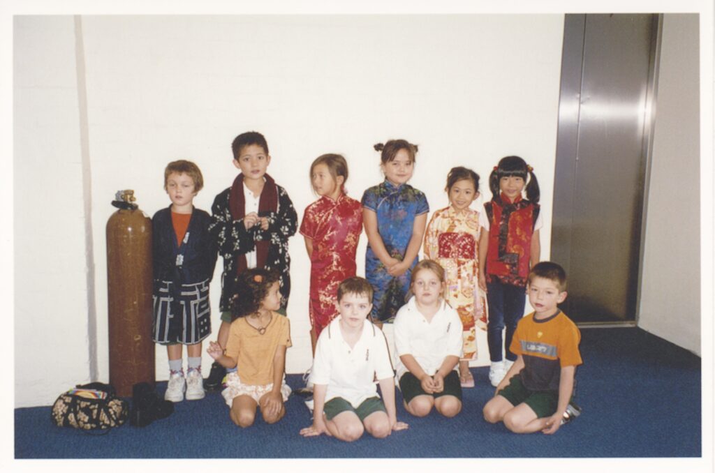 A group of nine young children pose indoors in harmony, some standing and some sitting. Reflecting diversity, several girls wear colourful traditional dresses whilst others are in casual clothes. A tall metal tank and a bag sit on the left side of the image.