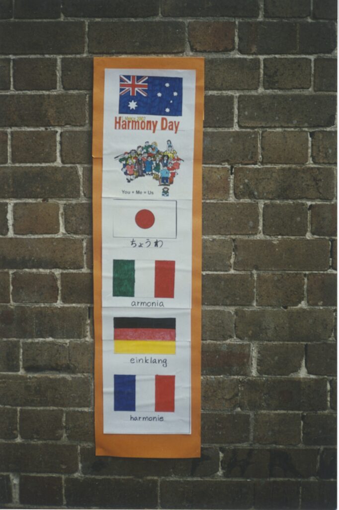 A Harmony Day poster on a brick wall shows the Australian flag, a cartoon group of diverse people, and the word Harmony in Japanese, Spanish, German, and French, each paired with its national flag.