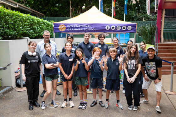 A group of students and adults from IGS pose and smile in front of a yellow tent labelled “International Grammar School” in an outdoor courtyard, some waving at the camera during the Waste Audit 2026 event.