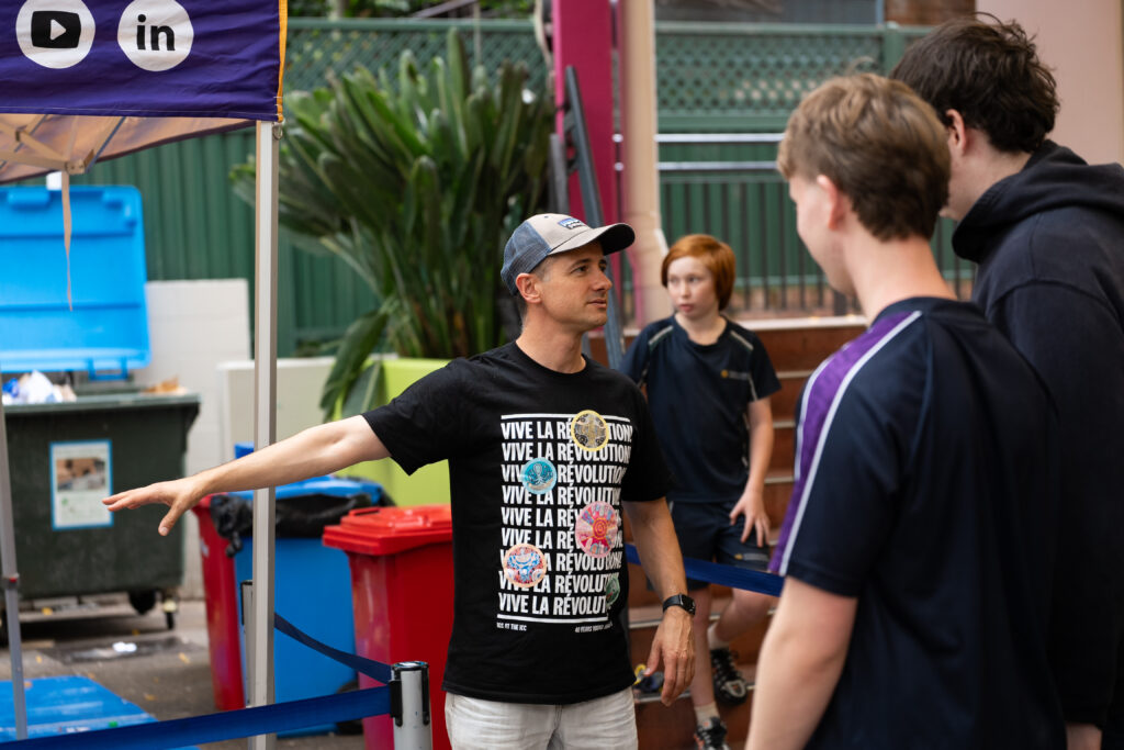 A man in a black Vive la Révolution t-shirt gestures whilst speaking to two young men outdoors during the IGS Waste Audit 2026, with recycling bins and a boy in sports kit visible in the background.