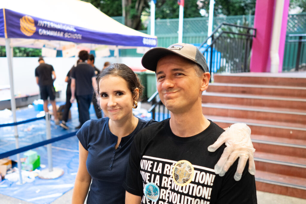 A woman and a man stand close together, smiling at the camera. The man wears a cap, glove, and bold IGS shirt. In the background, people are working under a marquee near a staircase during the Waste Audit 2026 event.
