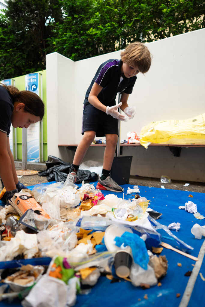 Two young people wearing gloves conduct a Waste Audit, sorting through a pile of assorted rubbish and recyclables on a blue tarpaulin outdoors, with greenery and a white wall in the background—an IGS initiative for 2026.