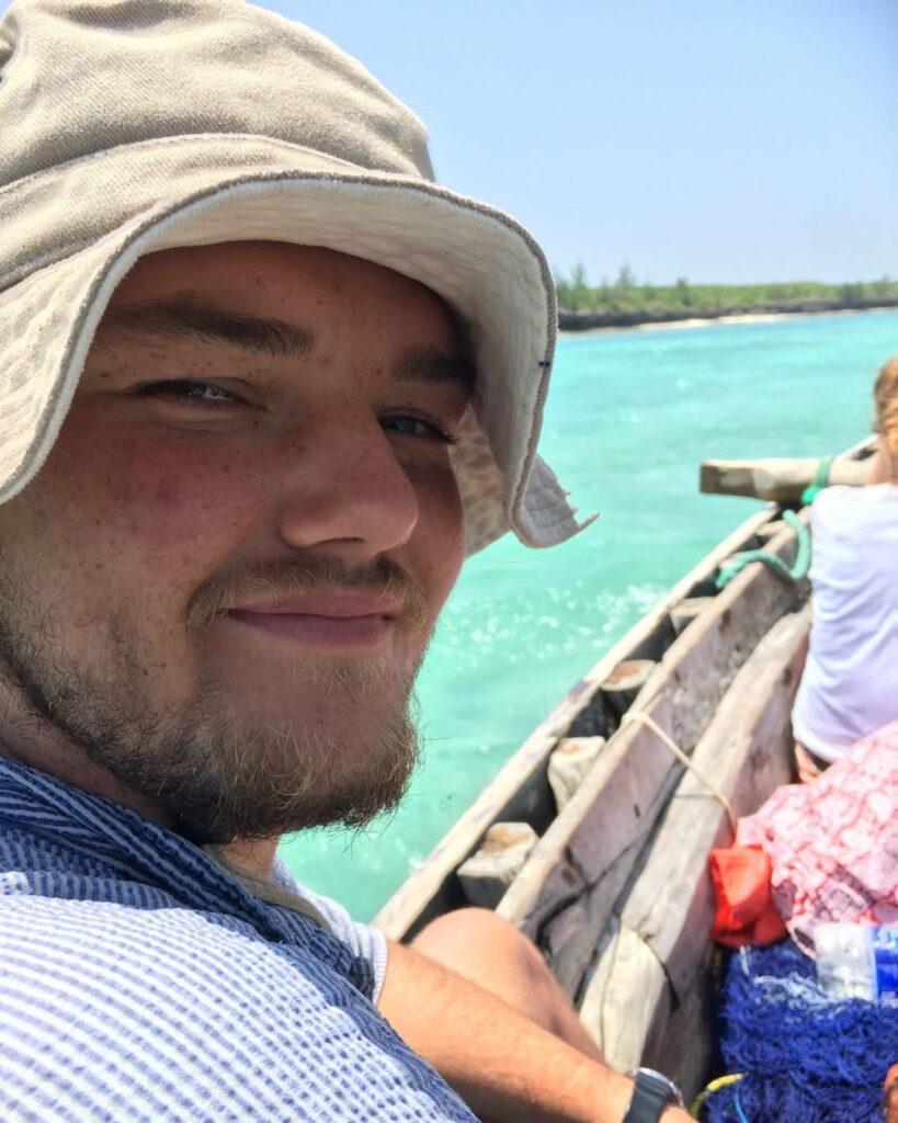 A person wearing a light-coloured sun hat sits smiling in a wooden boat on bright turquoise water, with another person and supplies visible, under a clear blue sky—enjoying the perfect #33 Tripleplay adventure.