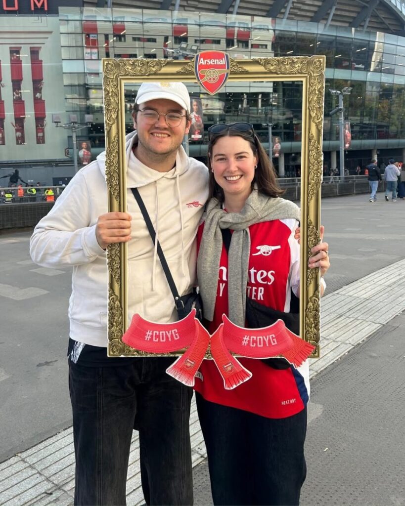 A smiling man and woman pose together outside a stadium, holding a large ornate frame with an Arsenal logo and #COYG scarves. The woman wears a red Arsenal shirt. Tripleplay spirit shines as #33 pride is on full display.