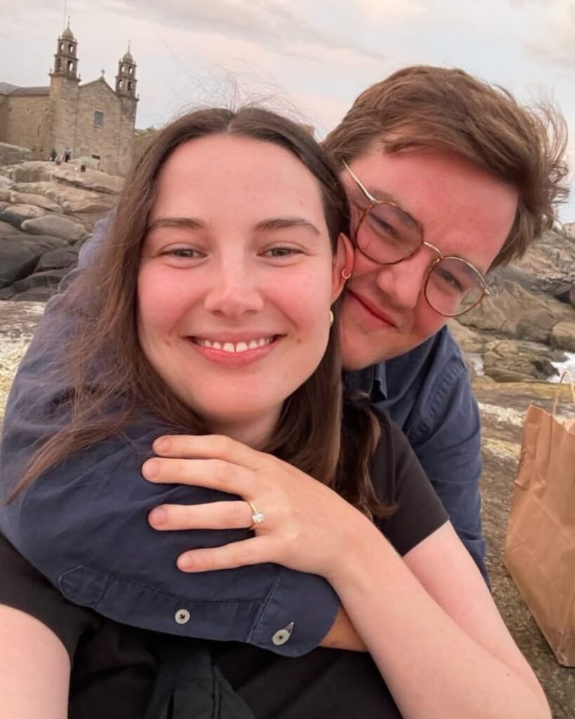 A smiling couple pose for a selfie on rocky terrain, the woman showing off her #33 engagement ring as the man hugs her from behind. A historic stone church stands in the background, capturing their Tripleplay moment outdoors.