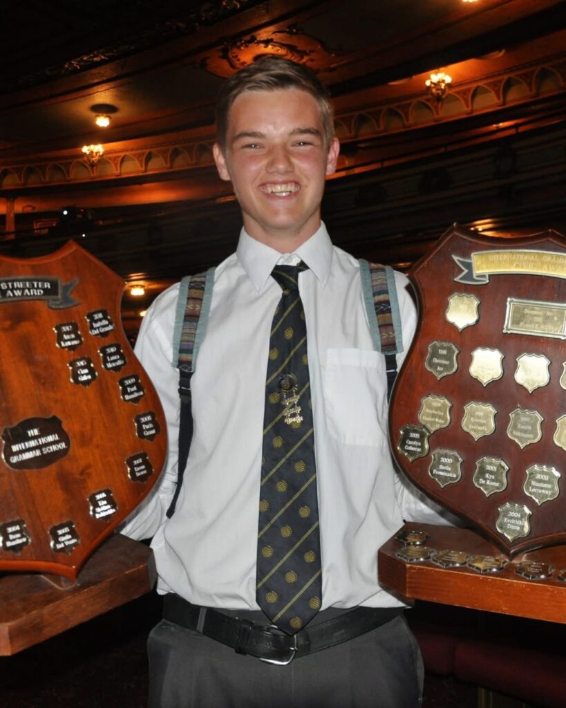 A young man wearing a shirt and tie smiles whilst holding two large wooden award plaques, including one for WITWAT, in a decorated indoor setting, possibly during the Tripleplay No. 33 ceremony or event.