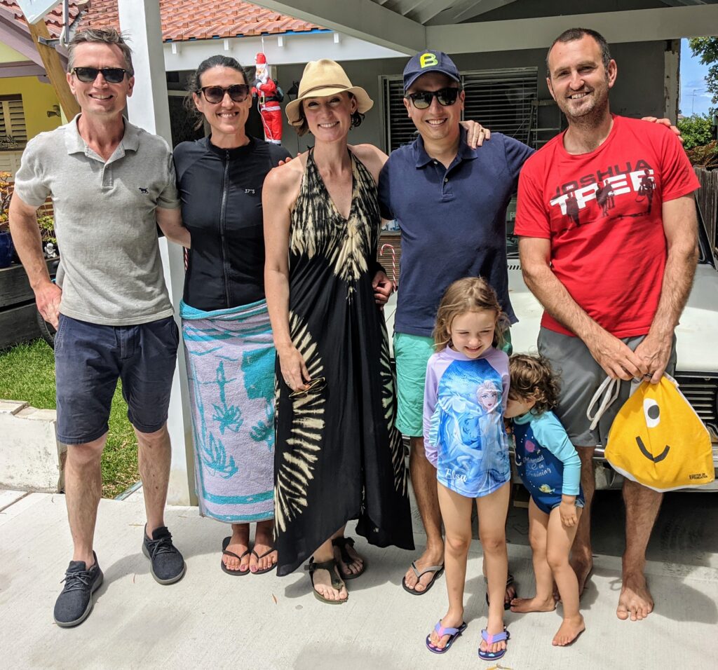 Six adults and two children pose and smile outdoors. One child wears a Frozen swimming costume, and an adult holds a yellow swim float. The Tripleplay group stands casually on a sunny patio near a white house, enjoying the day together.
