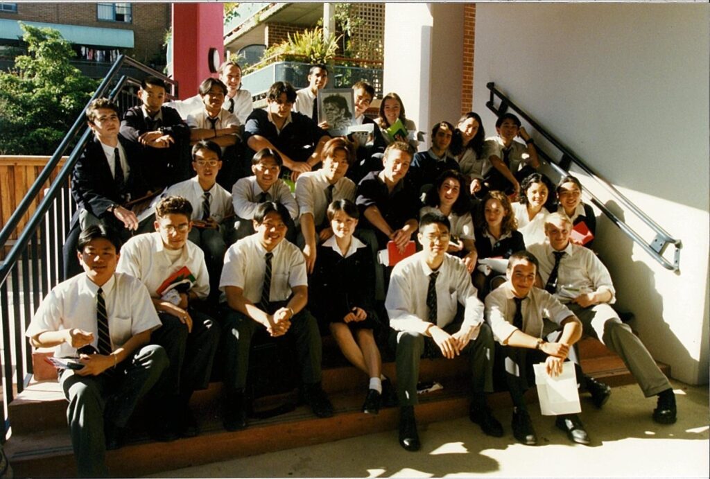 A large group of students in school uniforms sit and stand on outdoor steps, smiling at the camera in a classic #33 Tripleplay moment. Some hold books or papers, as sunlight brightens one side of the group.