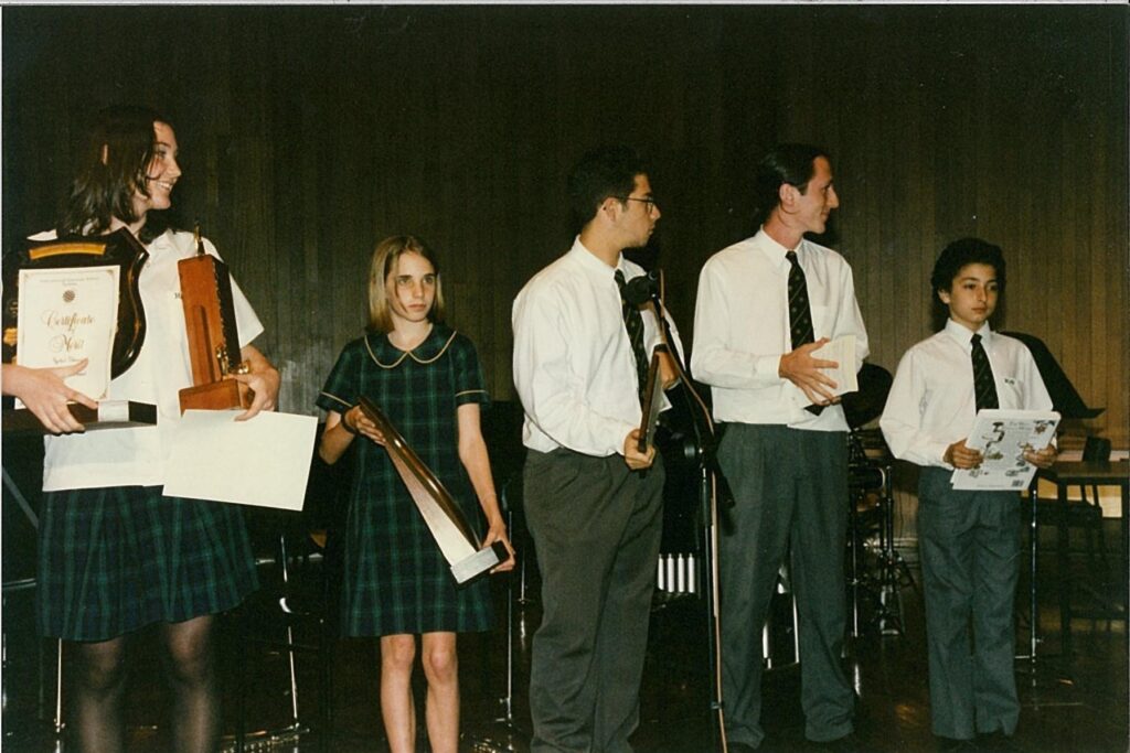 Five students stand on stage holding awards and certificates, dressed in school uniforms. Three wear green check, two are in white shirts and ties. At the WITWAT school prize-giving ceremony, they pose confidently against a wooden backdrop.