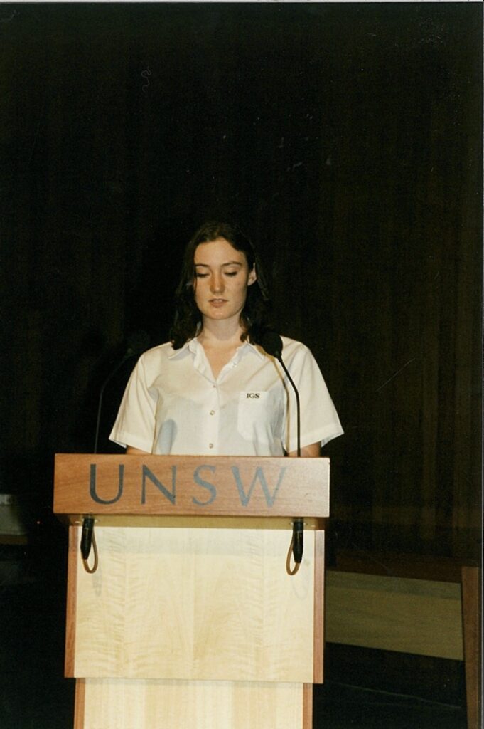 A young woman stands behind a lectern labelled UNSW, presenting with confidence. She is wearing a light-coloured short-sleeve shirt and has shoulder-length brown hair. The background is dark and plain, highlighting the Tripleplay event atmosphere.