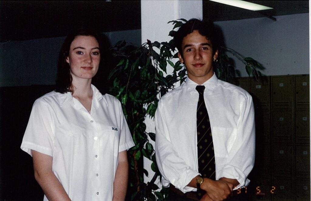 Two young people in white shirts stand indoors beside a tall plant and lockers. The person on the left has long hair; the one on the right, wearing a tie and watch, smiles slightly—capturing a candid moment behind the scenes of WITWAT episode 33.