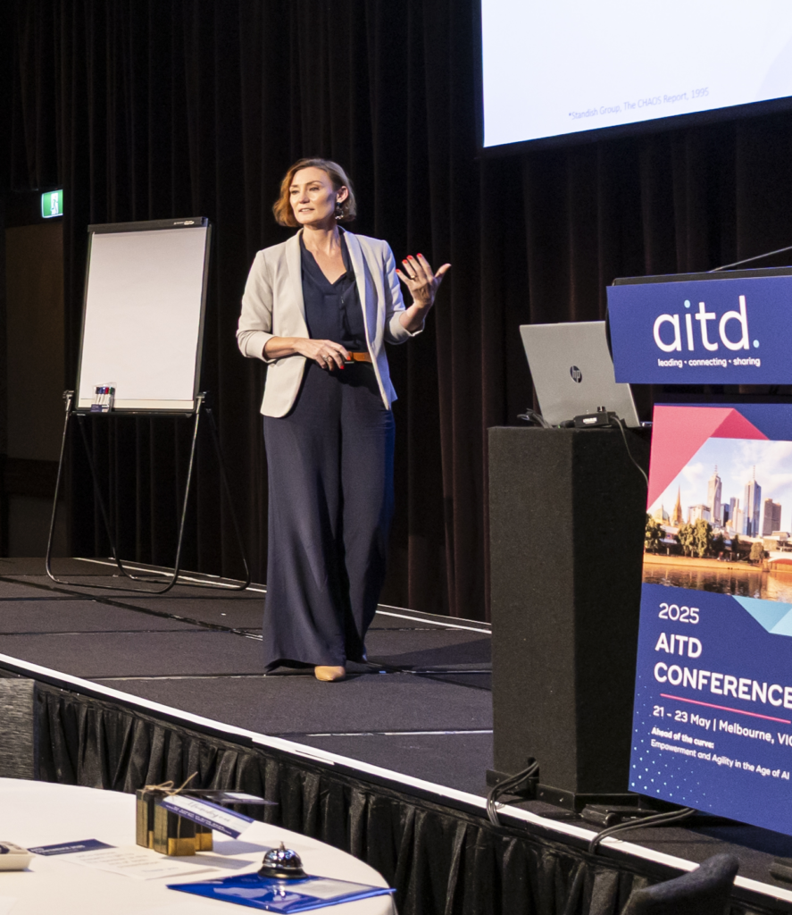 A woman in a blazer speaks on stage at the 2025 AITD Conference. Behind her are a flipchart and black curtain, with a lectern and #33 Tripleplay sign visible in the foreground.