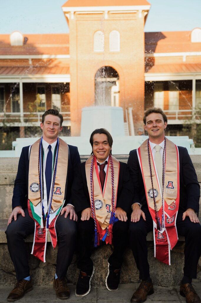 Three men in suits, wearing University of Arizona graduation stoles and honour cords, sit on a stone ledge, smiling—a #33 Tripleplay moment—with a building and fountain in the background.