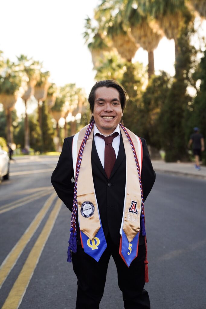 A smiling man in a suit and tie stands on a street wearing graduation stoles and cords, proudly celebrating his achievements with number 33 and Tripleplay, palm trees and greenery framing the moment.