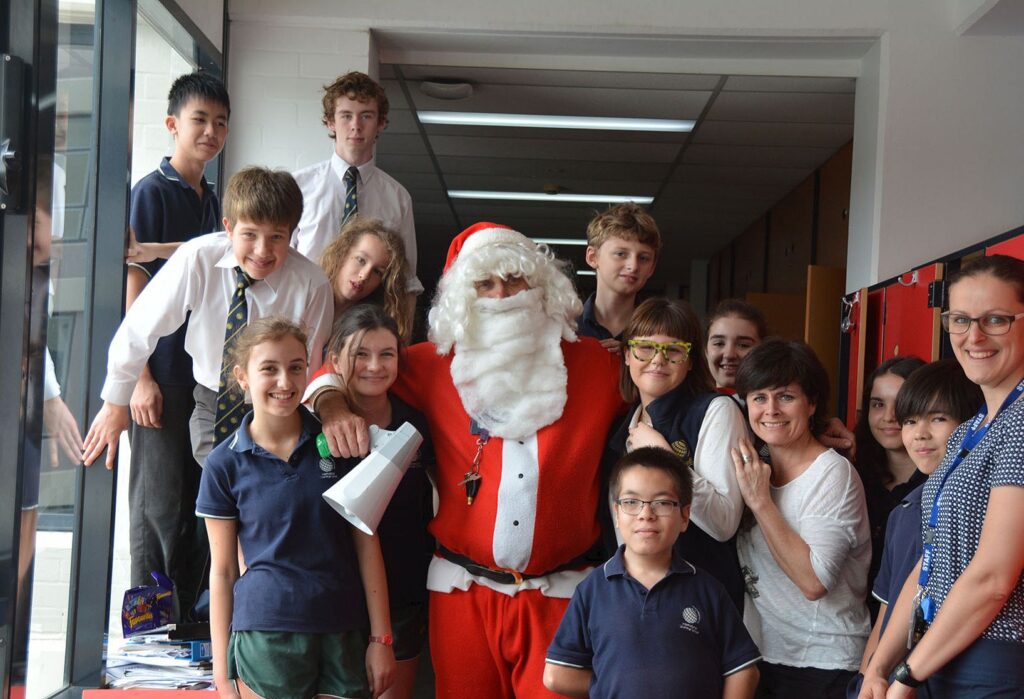 A group of students and teachers gather around a person dressed as Father Christmas in a school corridor, smiling and posing for a WITWAT photo, capturing the festive spirit with #33 and Tripleplay energy.