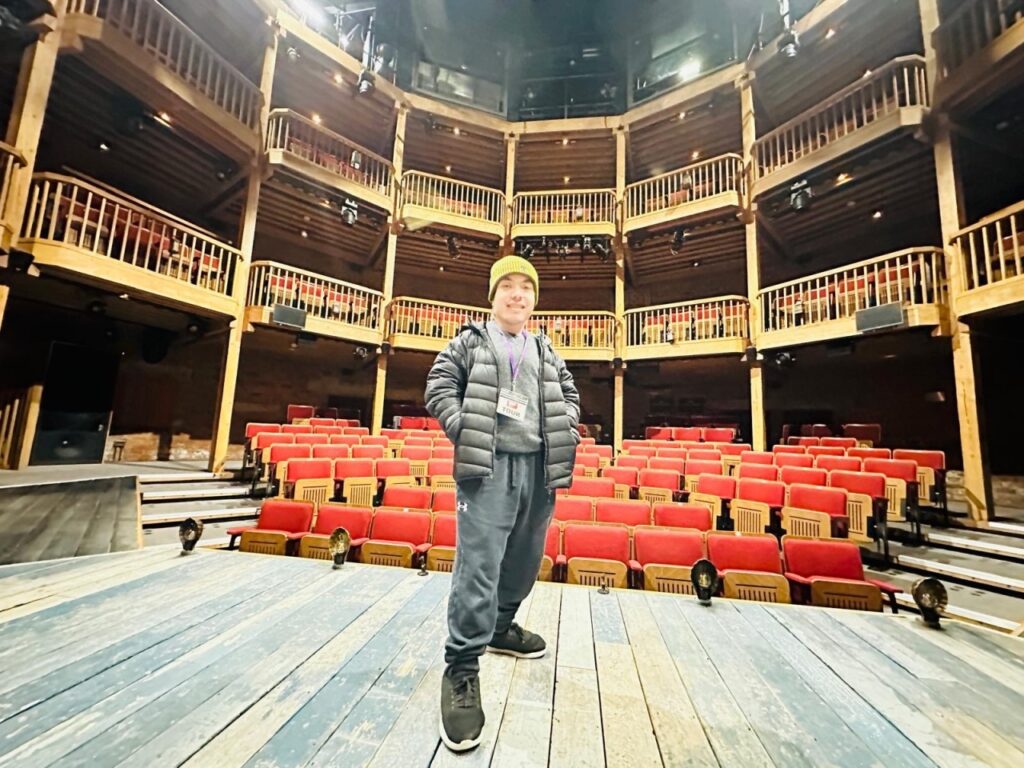 A person in a yellow hat and dark jacket stands confidently on a wooden stage, facing the camera. Behind them are rows of empty red theatre seats and a multi-level circular balcony in a warmly lit #33 Tripleplay theatre.