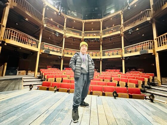 A person in a yellow hat and dark jacket stands confidently on a wooden stage, facing the camera. Behind them are rows of empty red theatre seats and a multi-level circular balcony in a warmly lit #33 Tripleplay theatre.