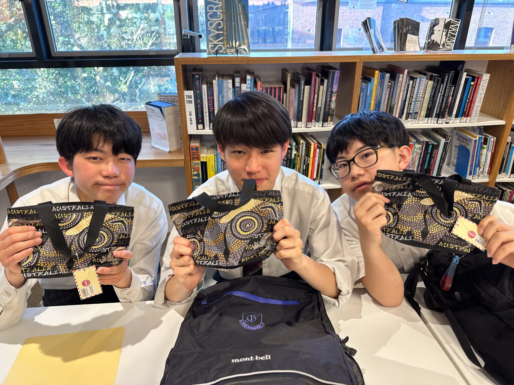 Three students in school uniforms sit at a table in a library, each holding up a colourful patterned pouch. Surrounded by bookcases and windows, they are building connections through shared language and creativity.