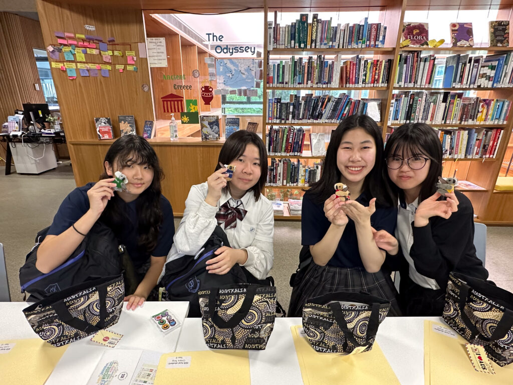 Four students sit at a table in a library, smiling and holding small figurines. Surrounded by bookshelves, they share patterned lunch bags, envelopes, and decorated boxes—building connections through language and laughter.