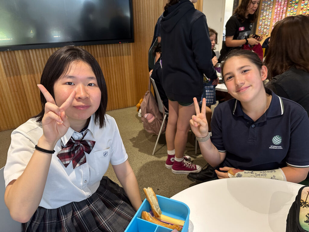 Two students sit at a table indoors, smiling and making peace signs. Sharing sandwiches from a blue lunchbox, they use language to build connections—one wears a checked skirt and tie, the other a navy polo shirt with a logo.