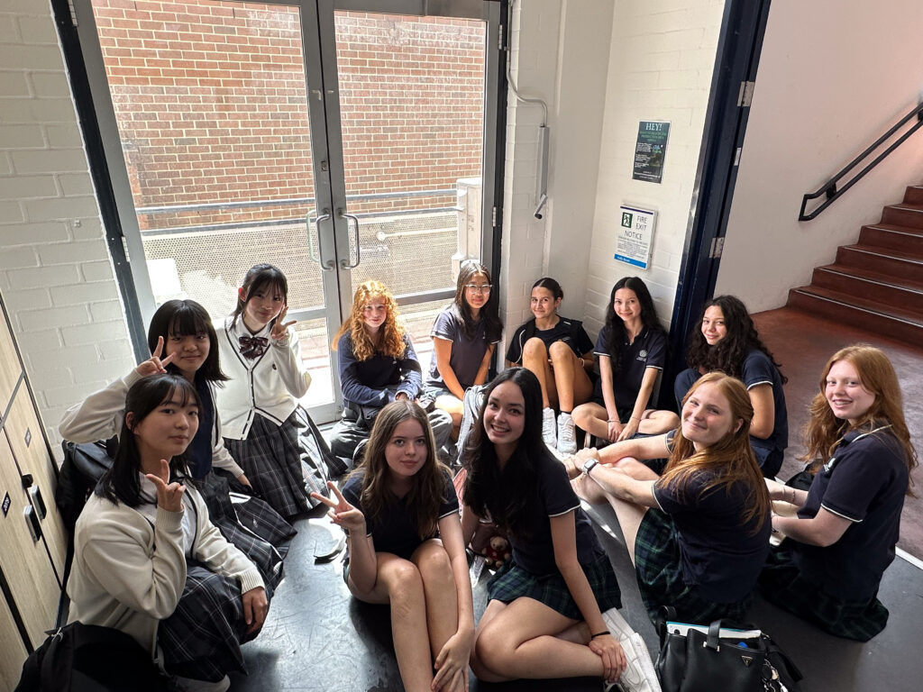 A group of twelve teenage girls in school uniforms sit in a circle near a glass door, smiling and making peace signs. Sunlight pours in as they enjoy building connections through shared language, with lockers and stairs visible behind them.