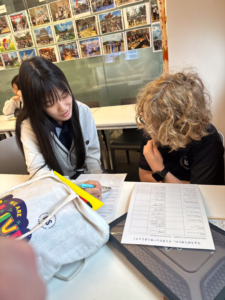 Two students sit at a desk, building connections as they work together on a language worksheet. One points with a highlighter, both focused on the paper. The classroom wall behind them is covered with colourful photos.