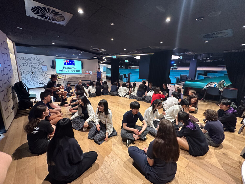 A group of students sit in small circles on a wooden floor indoors, building connections through lively discussions. A screen at the front reads “Favourite anime character.” The room features modern decor and bright lighting.