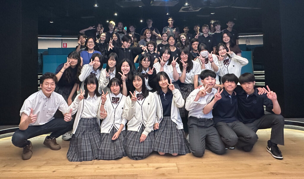 A large group of students in uniforms poses and smiles together indoors, some making peace signs. In a bright, modern room with tiered seating and a circular ceiling light overhead, they celebrate building connections through language.