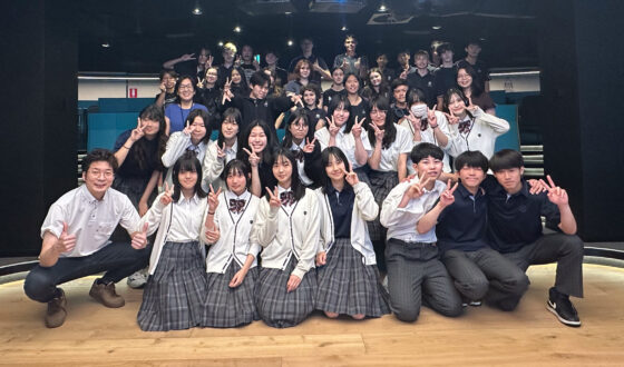 A large group of students in uniforms poses and smiles together indoors, some making peace signs. In a bright, modern room with tiered seating and a circular ceiling light overhead, they celebrate building connections through language.