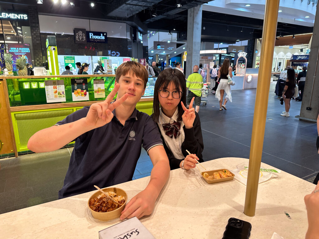 Two young people sit at a table in a shopping centre food court, eating from bowls and making peace signs towards the camera—building connections through shared moments and light-hearted communication as shoppers and food stalls bustle in the background.