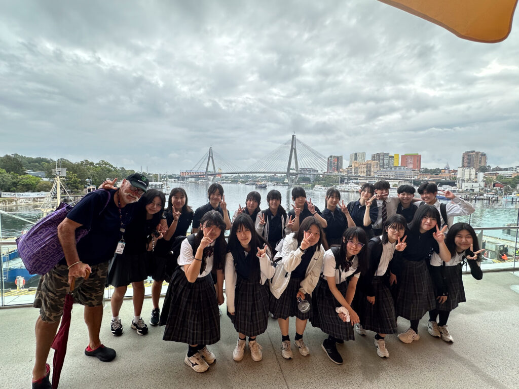 A group of students in school uniforms and a man in casual clothes pose on a balcony overlooking a harbour, building connections through language, with a modern cable-stayed bridge and city buildings under a cloudy sky in the background.