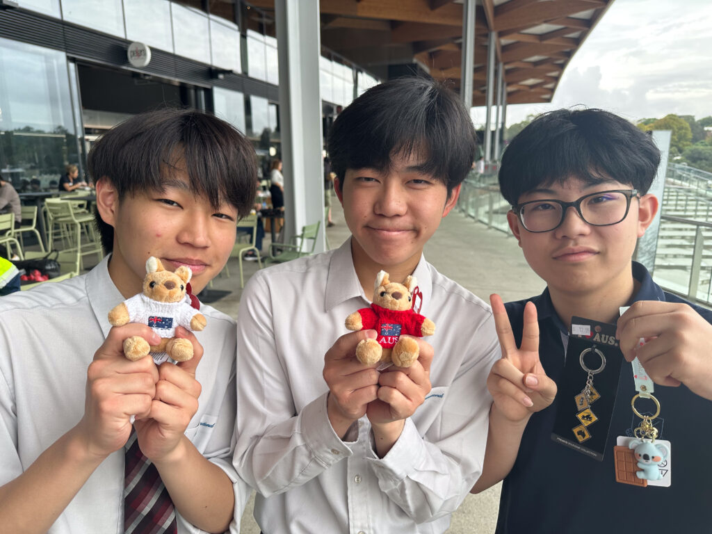 Three teenage boys smile and hold up small stuffed animal keyrings, making connections outside a modern building with glass walls and wooden beams. One boy makes a peace sign, highlighting the universal language of friendship.