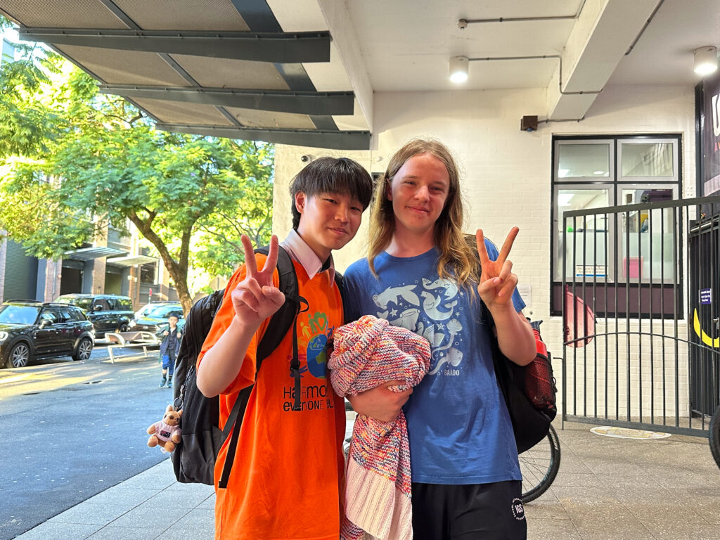 Two young people stand outdoors under a covered walkway, smiling and making peace signs. Through shared language, they're building connections. One wears an orange shirt and tie, the other a blue shirt holding a striped blanket. Trees and parked cars fill the background.