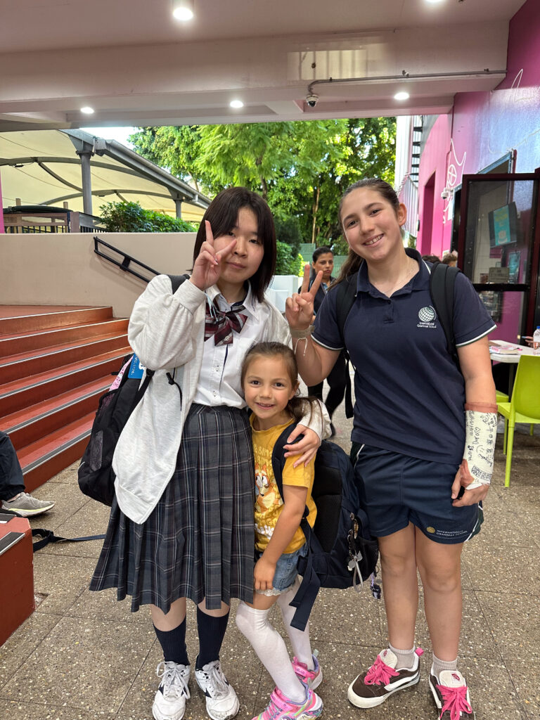 Three girls pose together, smiling and showing peace signs, building connections across styles—one in a checked school uniform, another in a sports kit with a plaster cast, and the youngest in a yellow shirt and pink shoes. Language bonds them all.