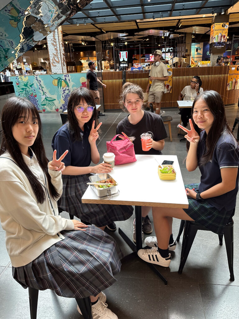 Four teenage girls sit around a table at a food court, smiling and making peace signs. Food and drinks are on the table. Their laughter shows how building connections goes beyond language, as others mingle in the colourful background.