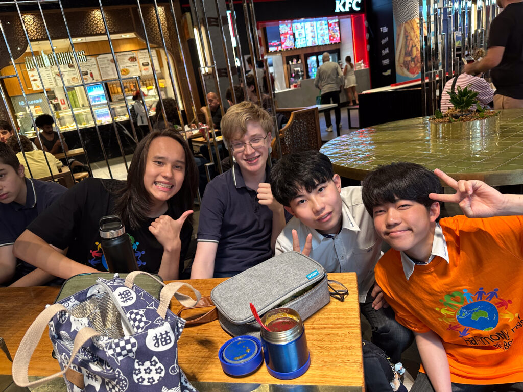 Four smiling teenagers sit at a table in a busy food court, building connections as they pose for a photo. Three make peace signs and one gives a thumbs up, with food containers on the table and a KFC restaurant in the background.