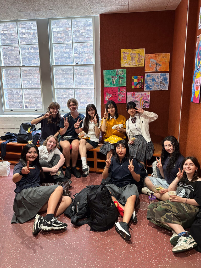 A group of students, some seated on benches and others on the floor, smile and make peace signs in a brightly lit room with large windows, colourful drawings, and a lively atmosphere for building connections through language.